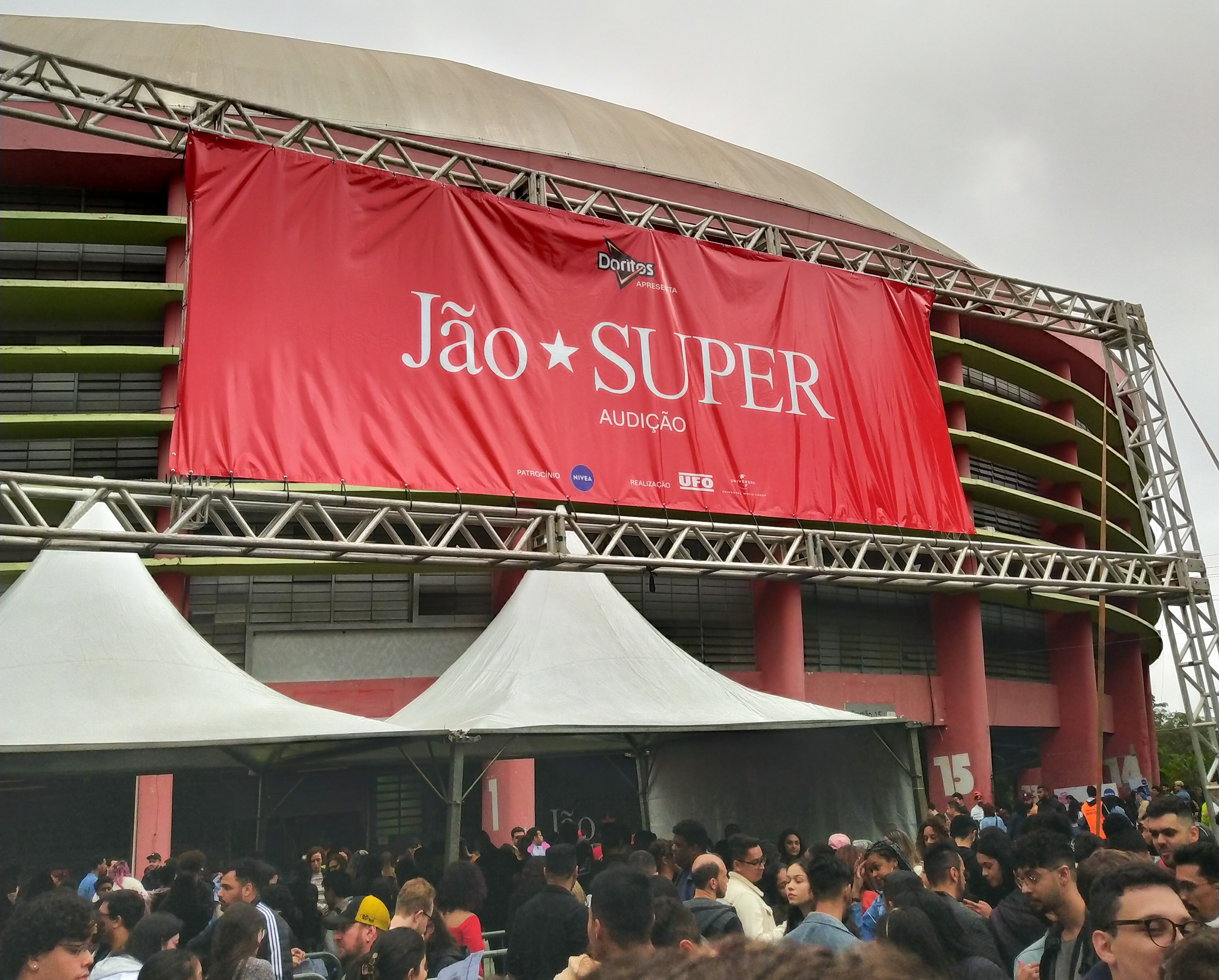 Na imagem há um banner vermelho com os dizeres "Jão Super, audição" à frente do Ginásio do Ibirapuera