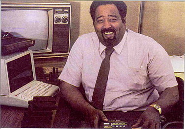 Homem negro adulto de camisa branca e gravata preta sorri para a câmera. Ele está próximo de aparelhos eletrônicos antigos - um computador e uma TV, além de um terceiro, de natureza desconhecida, que manuseia.