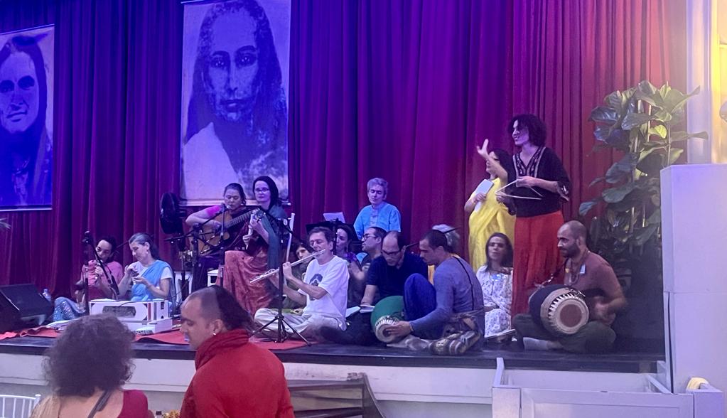 Varias pessoas de roupas coloridas sentadas no chão de um palco com cortinas vermelhas ao fundo, tocando instrumentos musicais orientais e cantando música tradicional indiana Kirtana