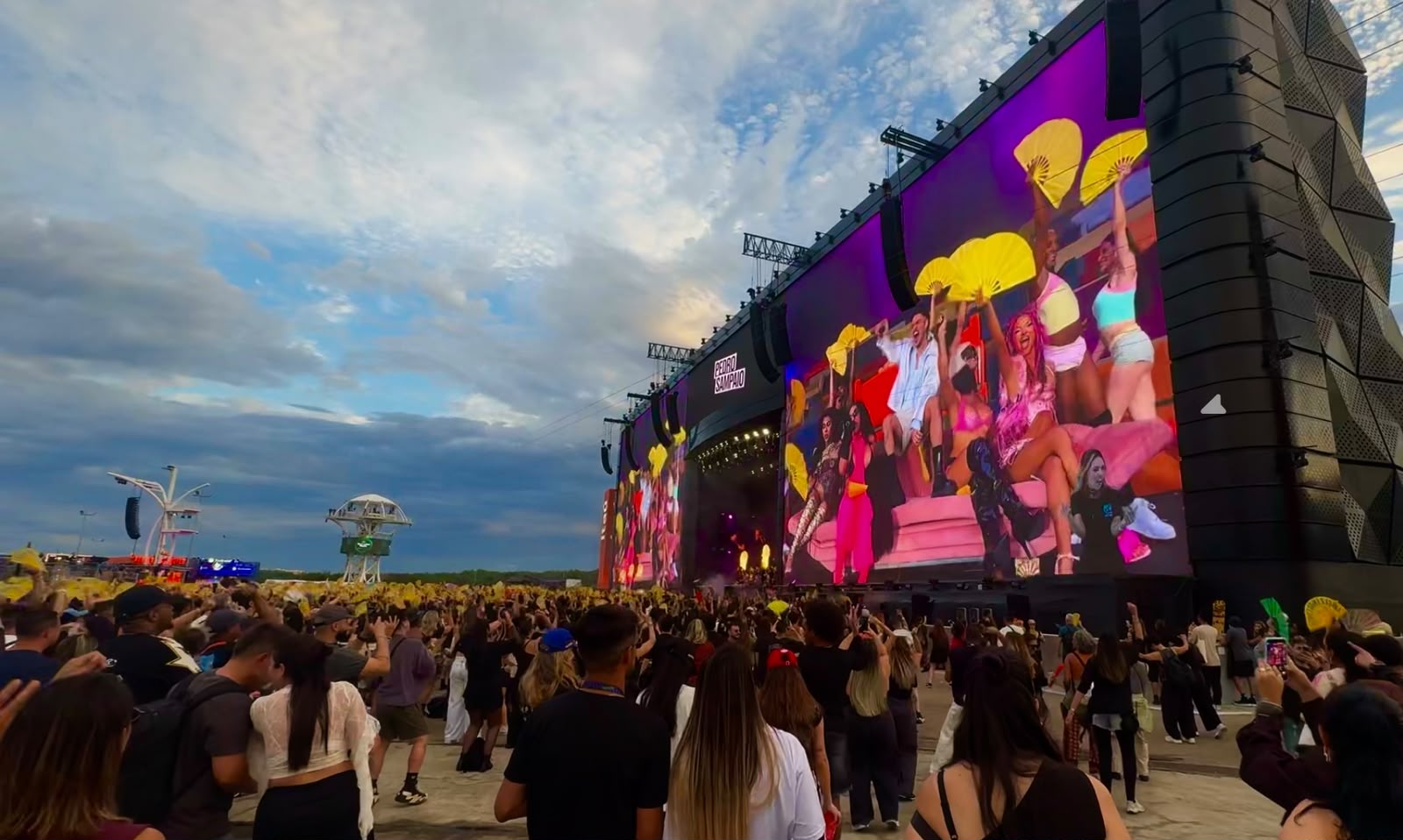 Público em um festival ao ar livre assistindo a uma apresentação exibida em grandes telões coloridos durante o entardecer.