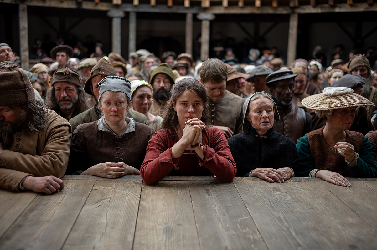 Cena do filme Hamnet: Agnes, personagem central da história, aparece sentada diante de uma multidão, com as mãos entrelaçadas perto do rosto e expressão apreensiva. Ela usa um vestido vermelho simples, enquanto pessoas ao seu redor vestem roupas de época e observam em silêncio, criando um clima de tensão.