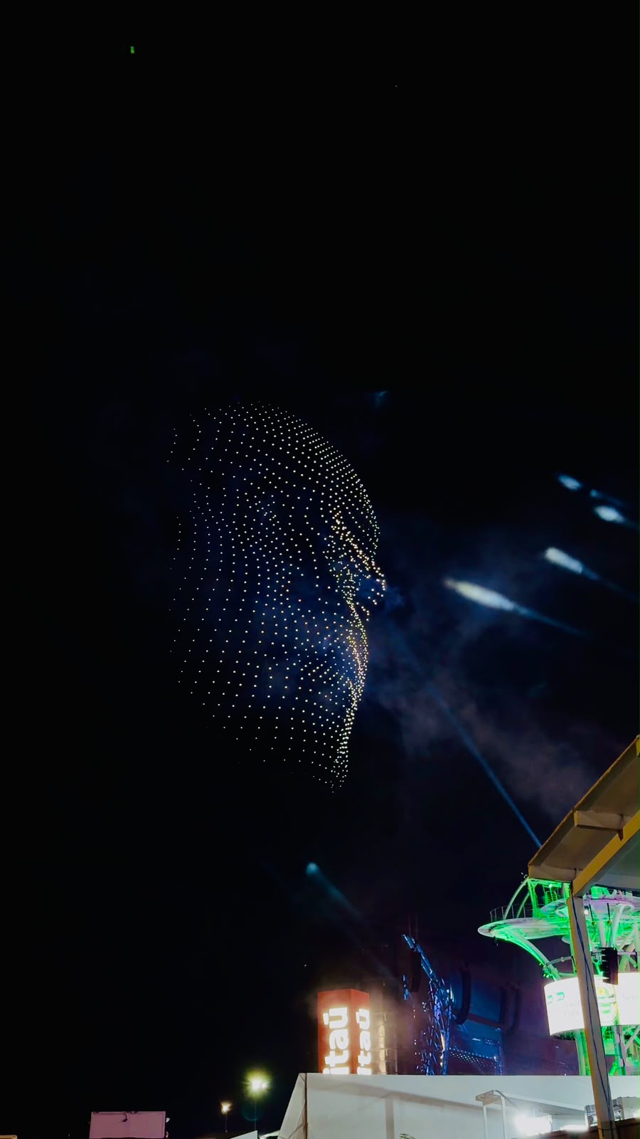 Formação de drones cria o desenho de um rosto no céu noturno durante um festival iluminado por lasers e luzes de palco.