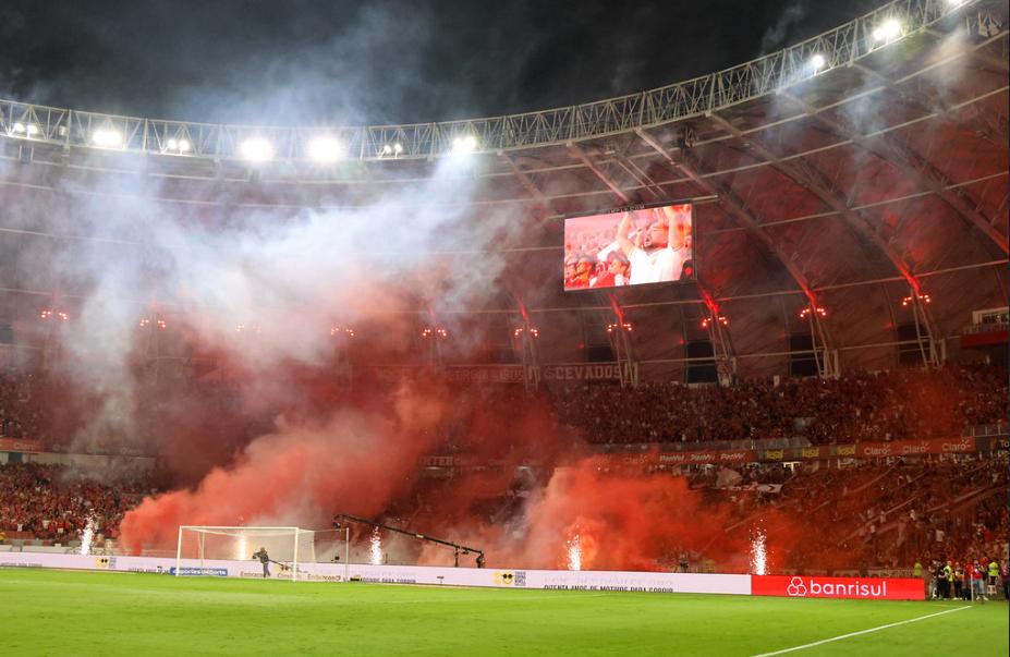A imagem mostra um setor da torcida do no Beira Rio. Há fumaça, oriundas de fogos de artificio na beira do campo, cobrindo parte da torcida.