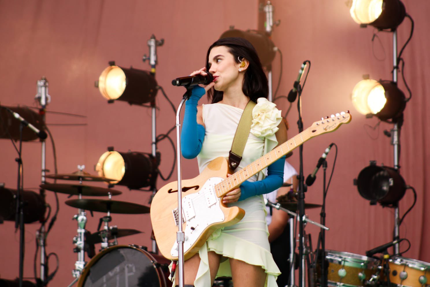 Em primeiro plano Manu Gavassi com uma guitarra laranja claro na mão vestindo um vestido curto branco e luvas azuis. No fundo da imagem há uma bateria, vários holofotes de cor amarelada e uma parede rosa.