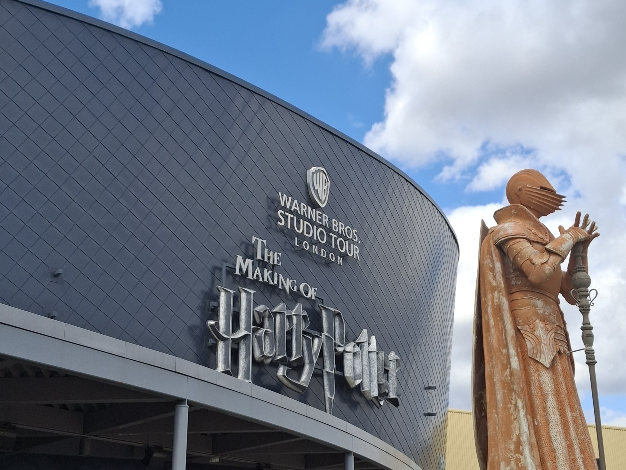 Fachada do prédio do Warner Bros. Studio Tour London – The Making of Harry Potter, com o logotipo da Warner Bros. e o título em letras grandes na parede curva do edifício. À direita, há uma grande estátua de um cavaleiro com armadura e capa, segurando uma espada.