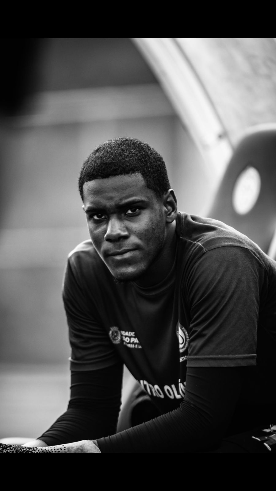 A fotografia mostra o atleta Cristian, sentado em um banco de reserva, olhando fixamente em direção à câmera. A foto está em preto e branco