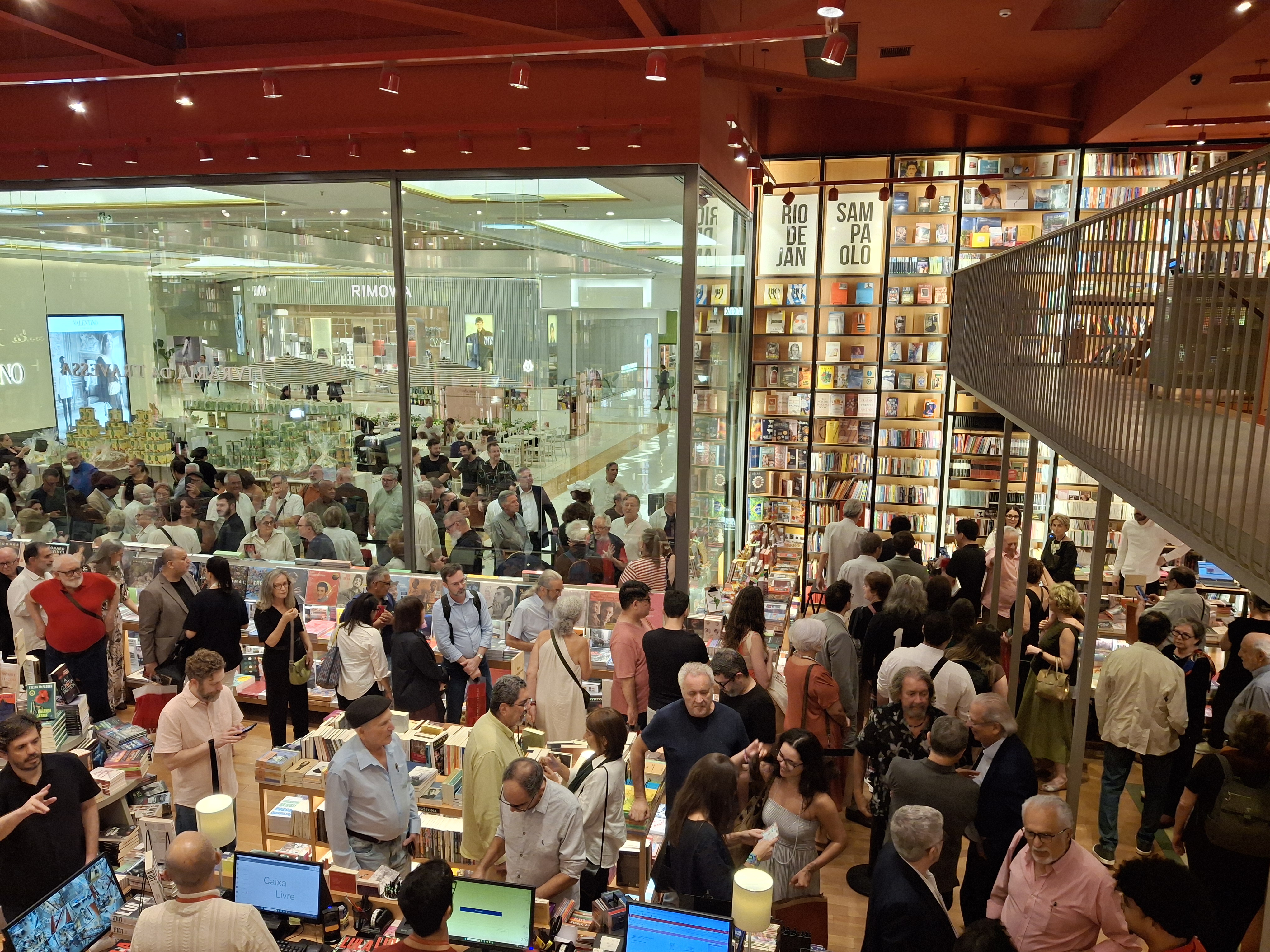 Texto alternativo: Grande público circula pelo interior da Livraria da Travessa, com pessoas reunidas entre mesas de livros e estantes altas. O espaço está cheio, sugerindo um evento ou lançamento literário, enquanto visitantes conversam e folheiam exemplares.