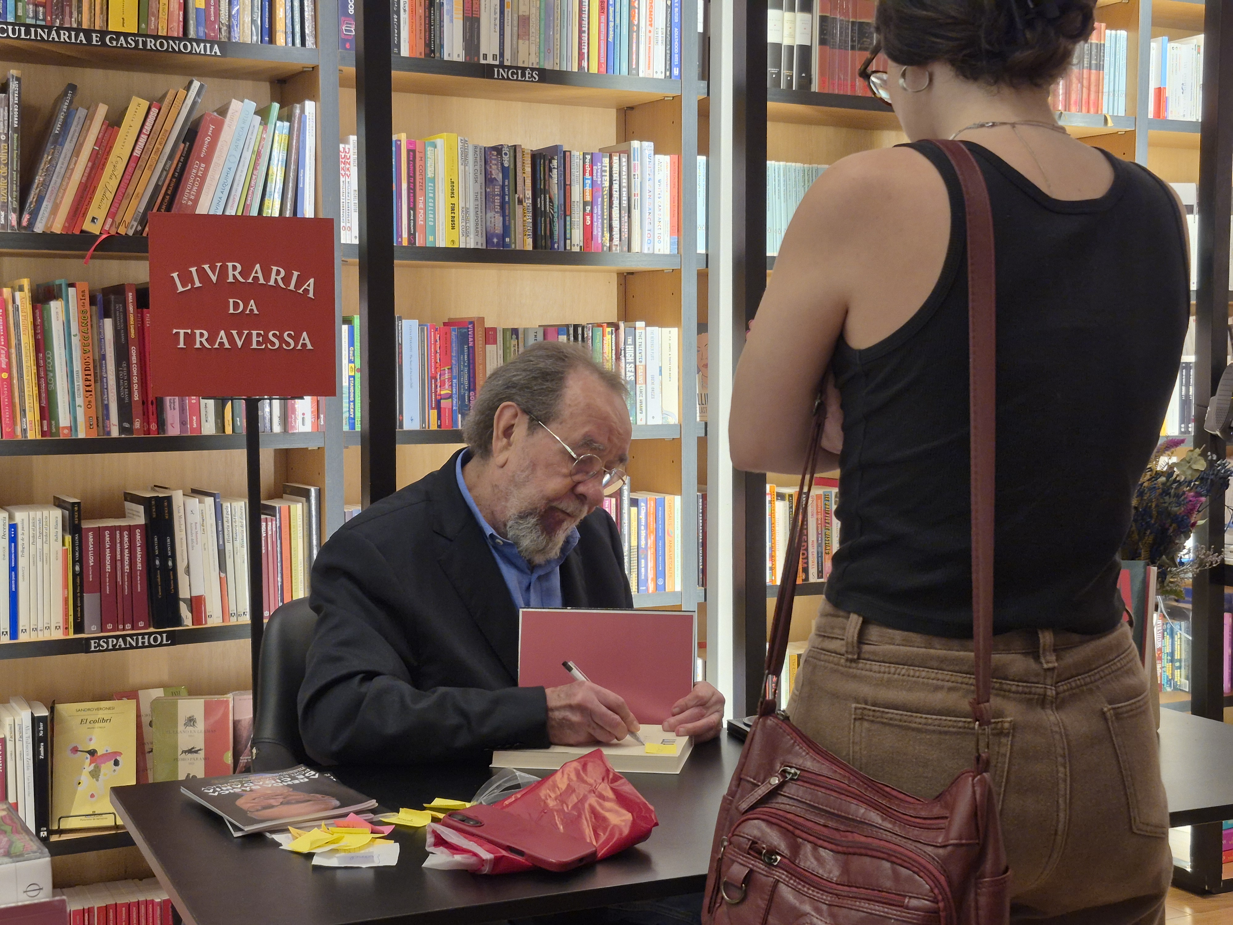 Homem autografa um livro para uma leitora à sua frente em uma mesa na Livraria da Travessa