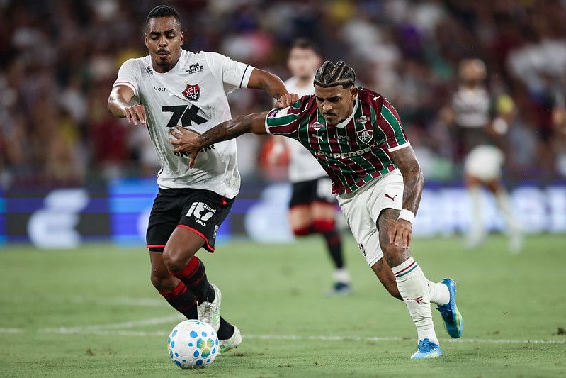 A imagem mostra uma disputa de bola entre um jogador do Fluminense e do Vitoria.