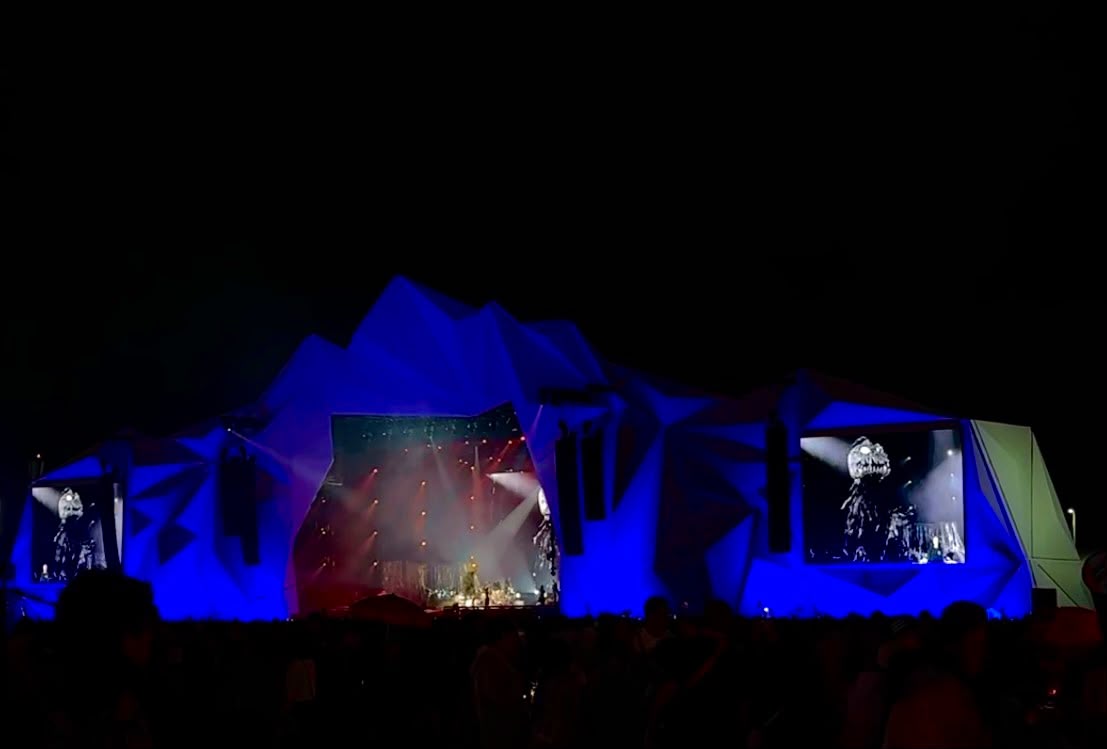 Palco de festival iluminado em azul durante a noite, com telões laterais exibindo o show e público reunido à frente.