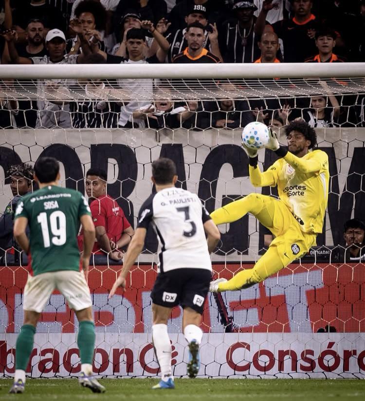 A imagem mostra o goleiro Hugo Souza, do Corinthians, em segundo plano, defendendo a bola. Gabriel Paulista, o Corinthians, e Sosa, do Palmeiras, olhando para o goleiro.