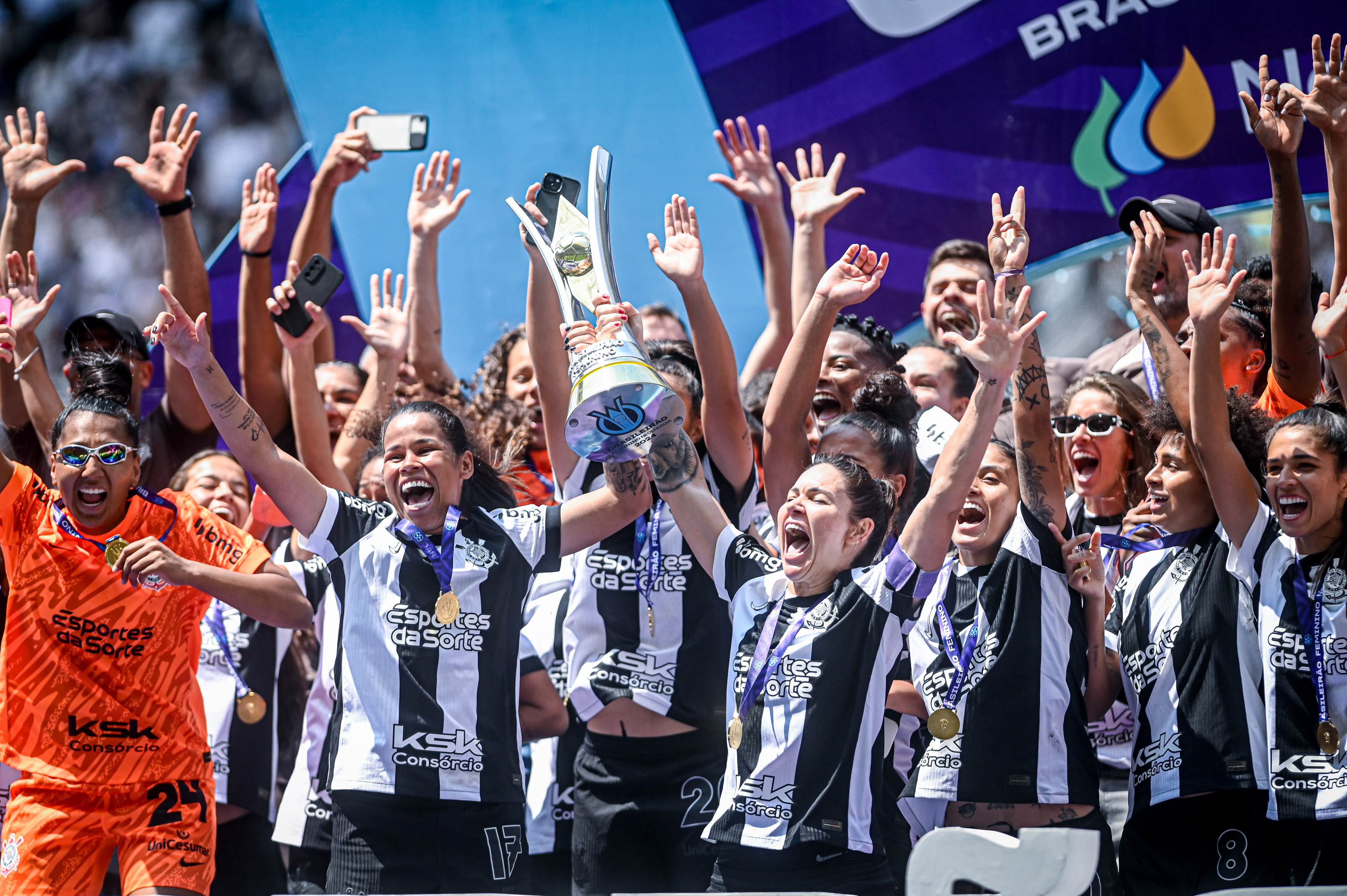 As capitãs do time, Gabi Zanotti e Vic Albuquerque, erguem a taça de campeãs brasileiras e as atletas comemoram.