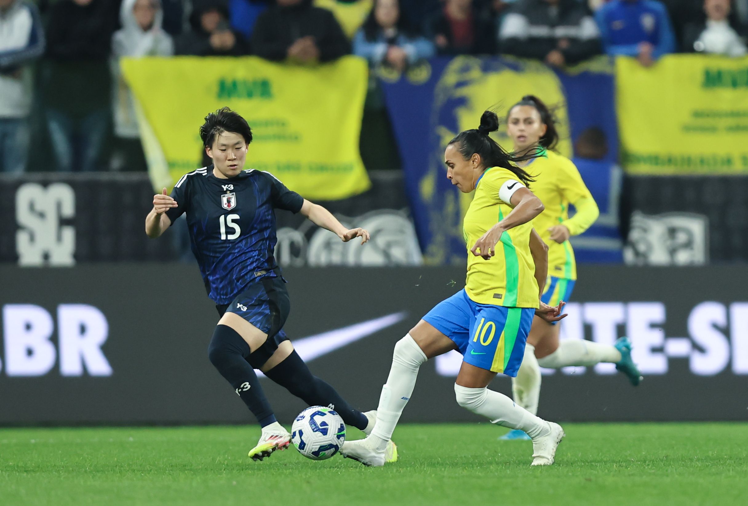 A imagem mostra a jogadora Marta, à direita, com o pé direito em direção a bola. Tentando interceptar a Marta, está a jogadora japonesa Fujino. Um pouco atrás de Marta está a brasileira Mariza. Ao fundo, há uma parte da torcida com algumas bandeiras
