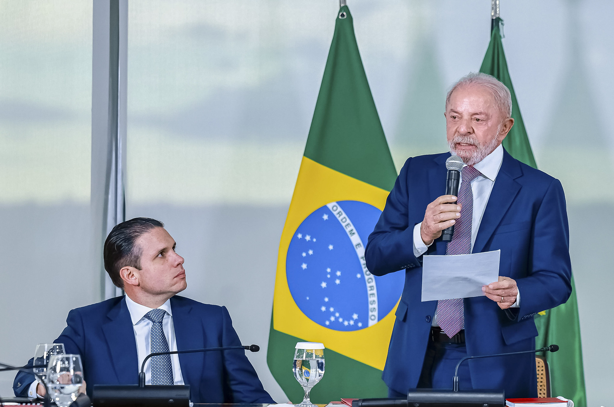 Luiz Inácio Lula da Silva aparece em pé, segurando um microfone e lendo um papel enquanto faz um discurso. Ao lado, Hugo Motta está sentado à mesa, olhando em sua direção. Ao fundo, aparecem bandeiras do Brasil, e sobre a mesa há microfones, copos de água e documentos.