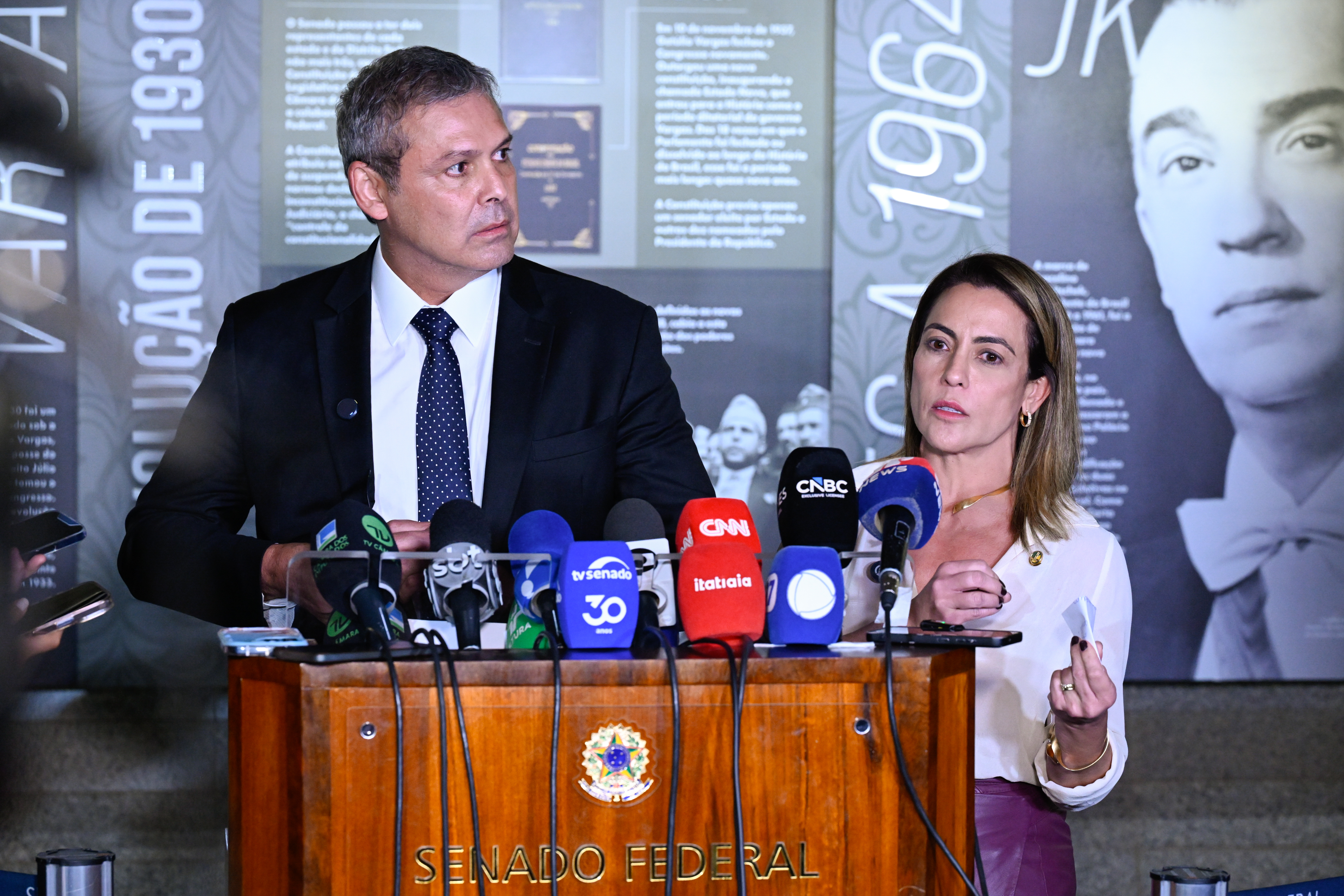Mulher e homem falam à imprensa em um púlpito do Senado Federal do Brasil, cercados por vários microfones de veículos de comunicação como CNN Brasil, TV Senado e CNBC. A mulher está falando enquanto segura um papel, e o homem observa ao lado. Ao fundo há um painel expositivo com imagens e textos históricos.