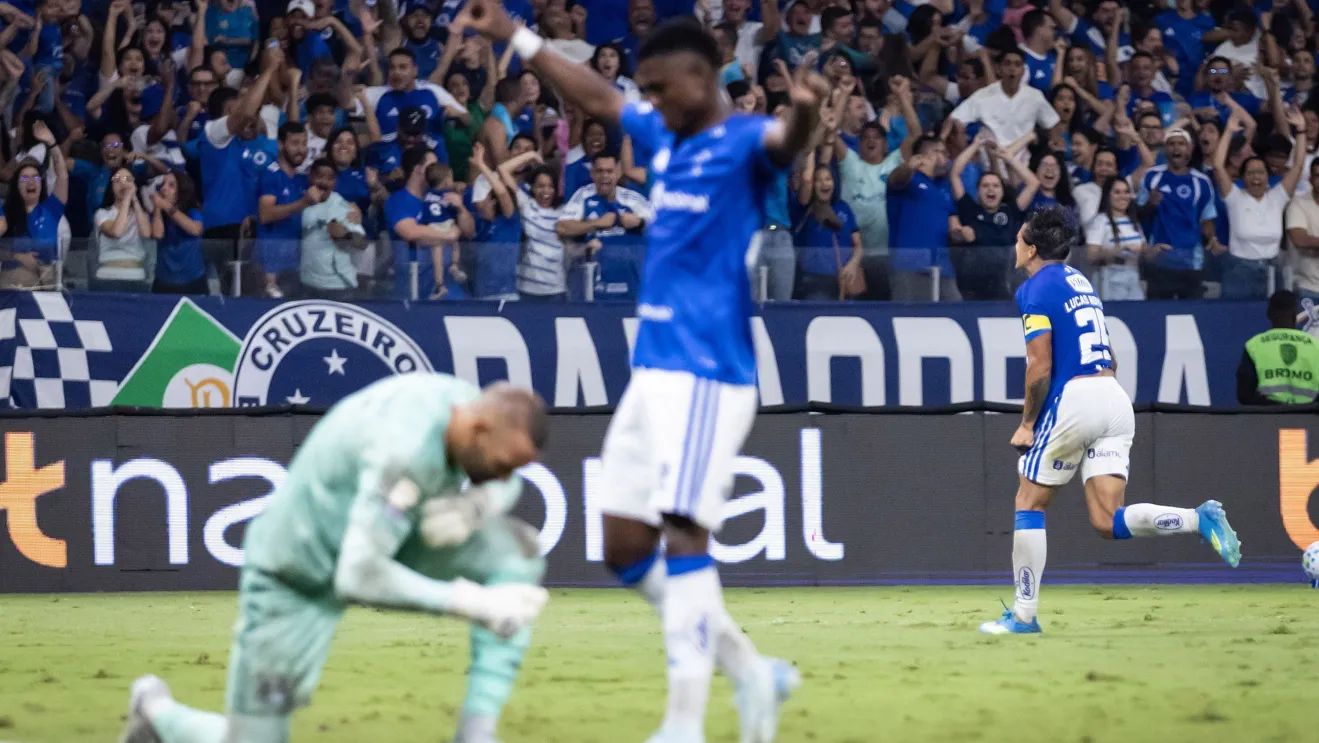 A imagem mostra o jogado Arroyo, do Cruzeiro, comemorando, enquanto Werton, goleiro do Grêmio está agachado. Ao fundo Lucas Romero, do Cruzeiro, também comemora em direção à torcida cruzeirense na arquibancada.