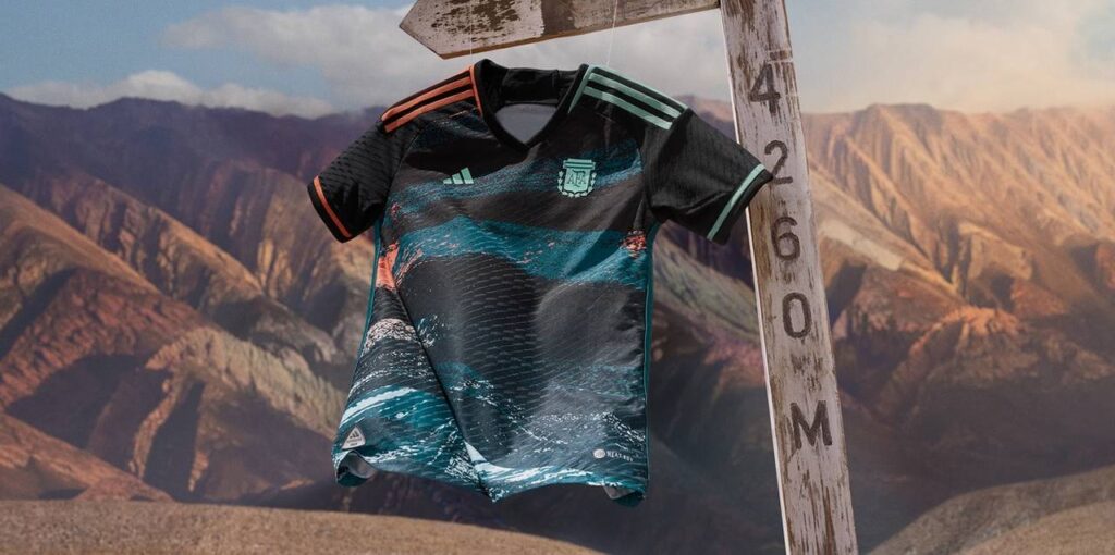 Camiseta da Adidas em azul e com linhas que lembram o relevo de montanhas. Foto de fundo: cordilheira na Argentina. 