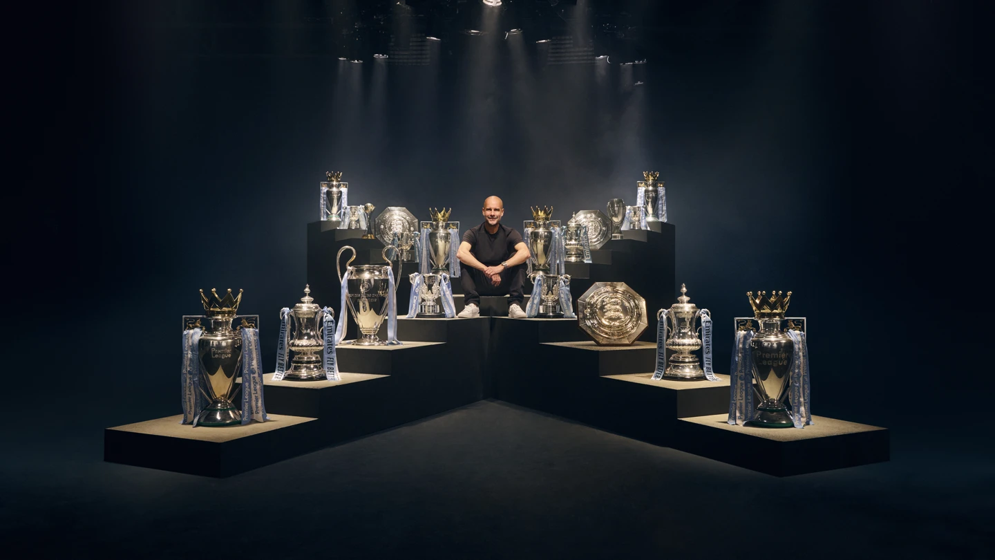A imagem mostra Pep Guardiola sentado com todos os troféus conquistados em volta
