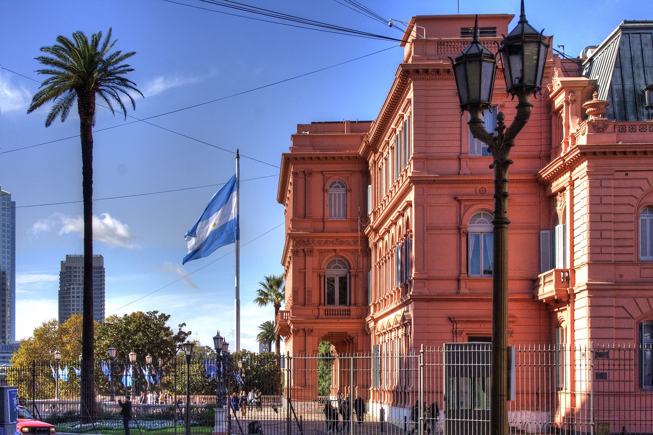 Edifício rosado, antigo, grande, sob céu azul e bandeira da Argentina. 