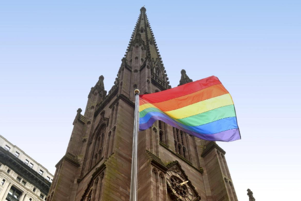 Atrás, uma parte da construção de uma igreja católica antiga e, á frente dela, uma bandeira da comunidade LGBTQIAPN+ erguida.