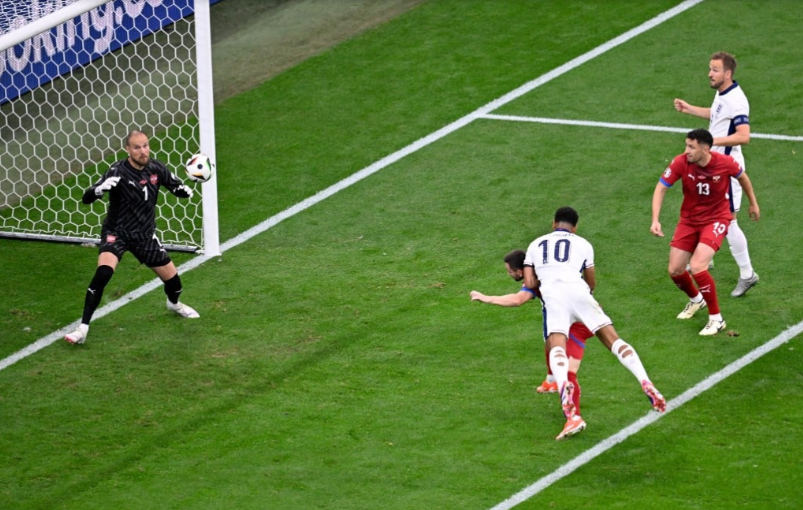 Na parte direita da imagem, dois jogadores da Inglaterra e dois jogadores da Sérvia estão na área pequena do campo de futebol. Enquanto na parte esquerda, o goleiro tenta defender a bola que um deles cabeceou.