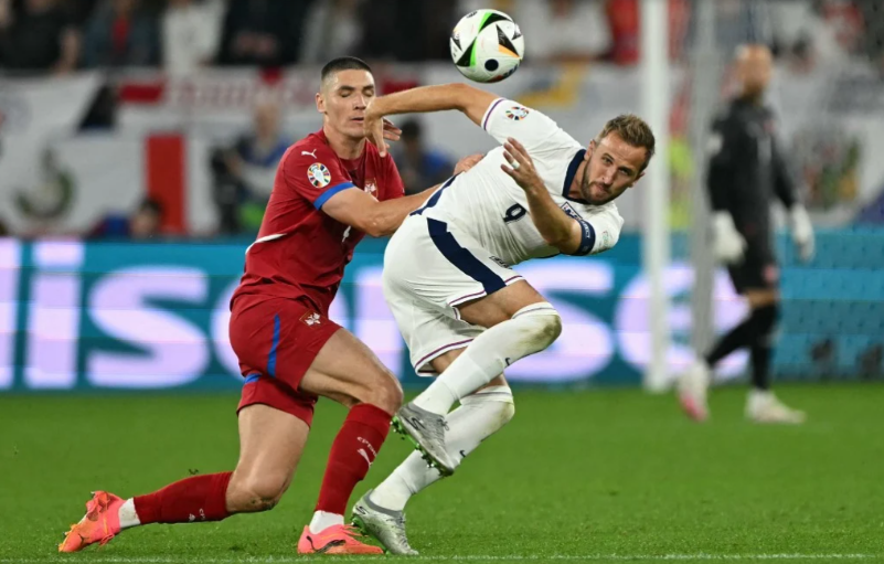 Um jogador da Sérvia, do lado esquerdo, disputa a bola com um jogador da Inglaterra, do lado direito, em um campo de futebol.