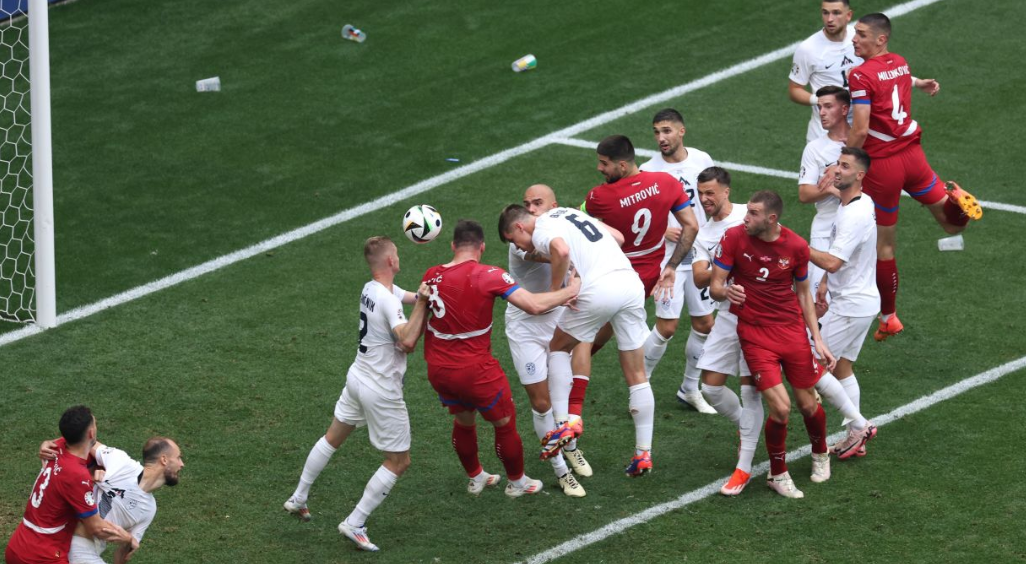 Jogadores da Eslovênia e Sérvia juntos na pequena área da seleção eslovena, pulando e tentando cabecear a bola em direção ao gol.