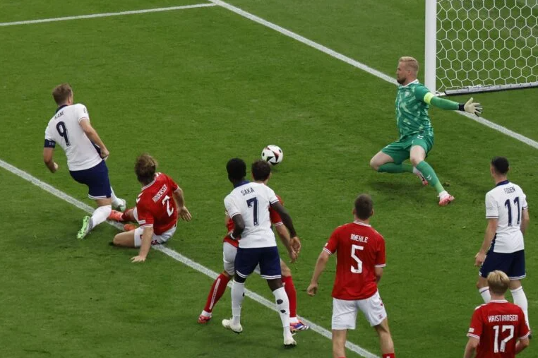 Jogadores da Inglaterra e Dinamarca próximos a pequena área dinamarquesa. Um deles, Harry Kane, chuta a bola em direção ao goleiro, na parte de baixo da esquerda do gol, enquanto o goleiro falha em pegar.