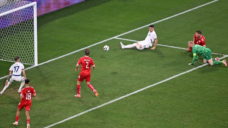 Dois jogadores da Sérvia em frente ao gol, três jogadores da Dinamarca logo atrás deles e o goleiro dinamarques caido ao lado dos jogadores do seu time, todos observam a bola entrar no gol.