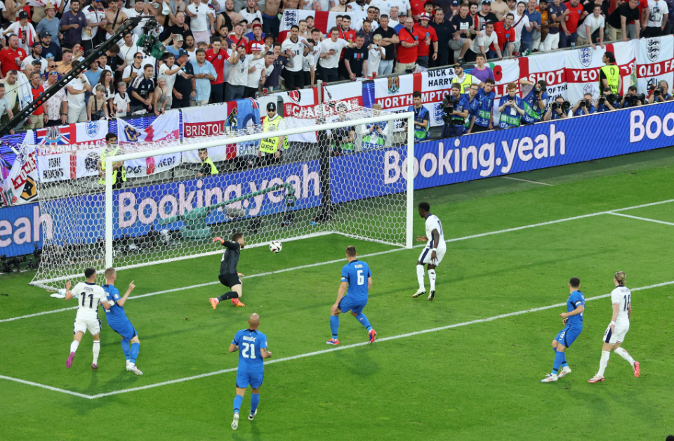 Jogadores ingleses e eslovenos próximos a pequena área inglesa, observando o goleiro inglês falhar na defesa do gol adversário.