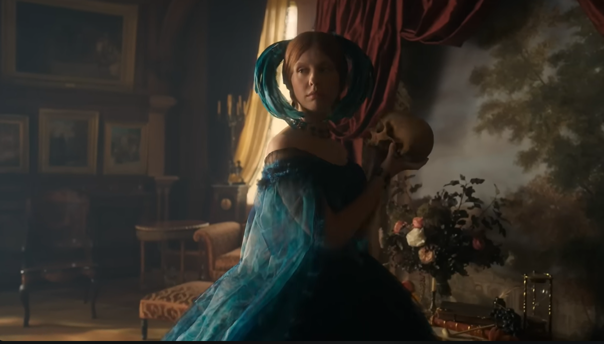 Foto: Personagem Elizabeth segura um crânio enquanto usa vestido verde com adereço de penas na cabeça. / Fonte: Netflix.