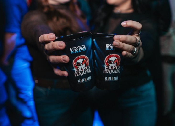 Foco em dois copos sendo brindados por mulheres. Os copos possuem logo e estética que remete ao evento de Itajubá.