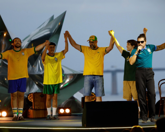 Atores de diferentes idades vestem camisas da Seleção Brasileira. Eles seguram a mão um do outro e as levantam em tom de agradecimento. Um cantor veste a camisa azul do time, e se apresenta para a plateia.