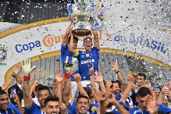 Homens com camisa azul e calção branco erguendo uma taça. Ao fundo, muito papel picado cai sobre eles