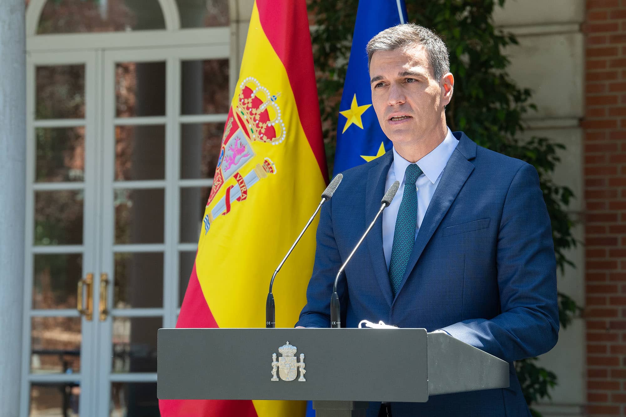 Primeiro- Ministro da Espanha, Pedro Sánchez, discursando em um púlpito. Ao fundo, estão uma bandeira da Espanha e outra da União Europeia.
