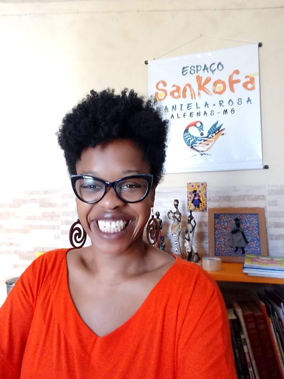 A imagem mostra Daniela Rosa, mulher negra, atrás de um painel escrito "Espaço Sankofa", local onde ela atende como doula. além de ter uma mesa ao fundo com esculturas e quadros da cultura afrodescendente