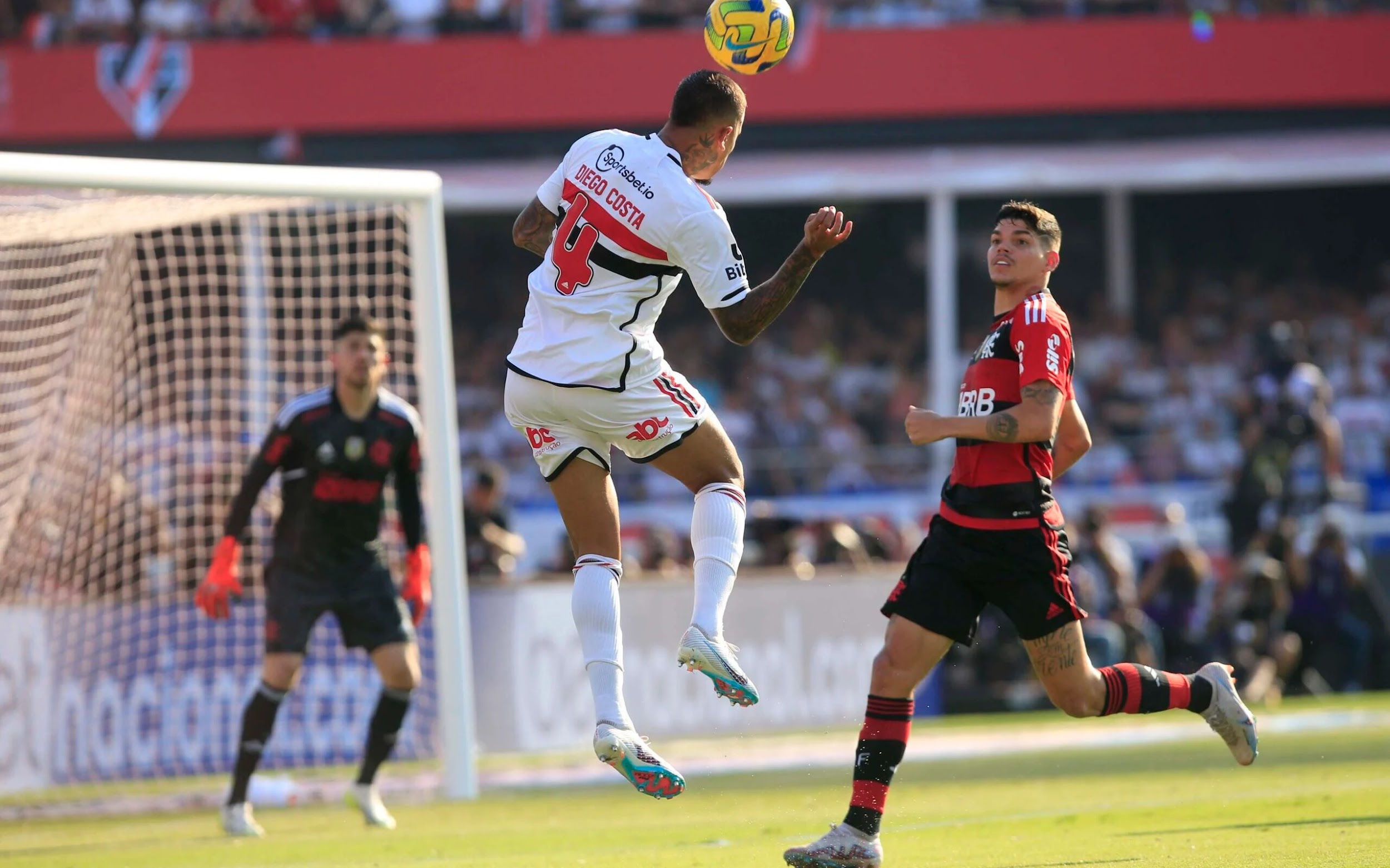 Homem com camisa, calção, meião e chuteiras brancas salta e cabeceia a bola. Na imagem, também tem um homem, com camisa listrada preta e vermelha, calção preto, meião listrado preto e vermelho e chuteira branca. Ao fundo, homem com camisa, calção e meião preto, com luvas vermelhas.