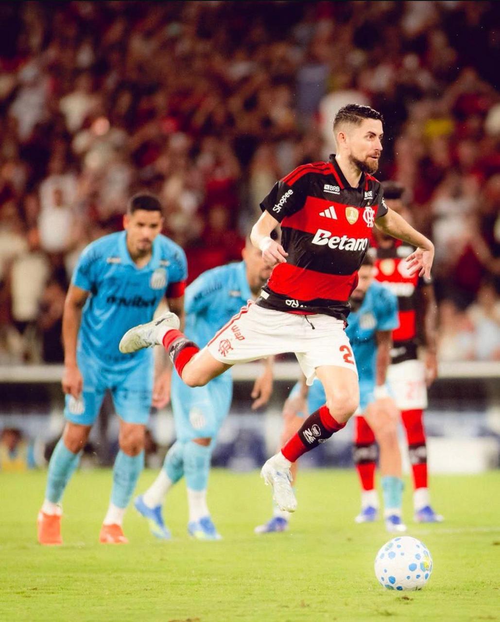 A imagem mostra o jogador Jorginho no ar antes de chutar a bola parada na marca do pênalti. Ao fundo, há jogadores do Santos aguardando a cobrança