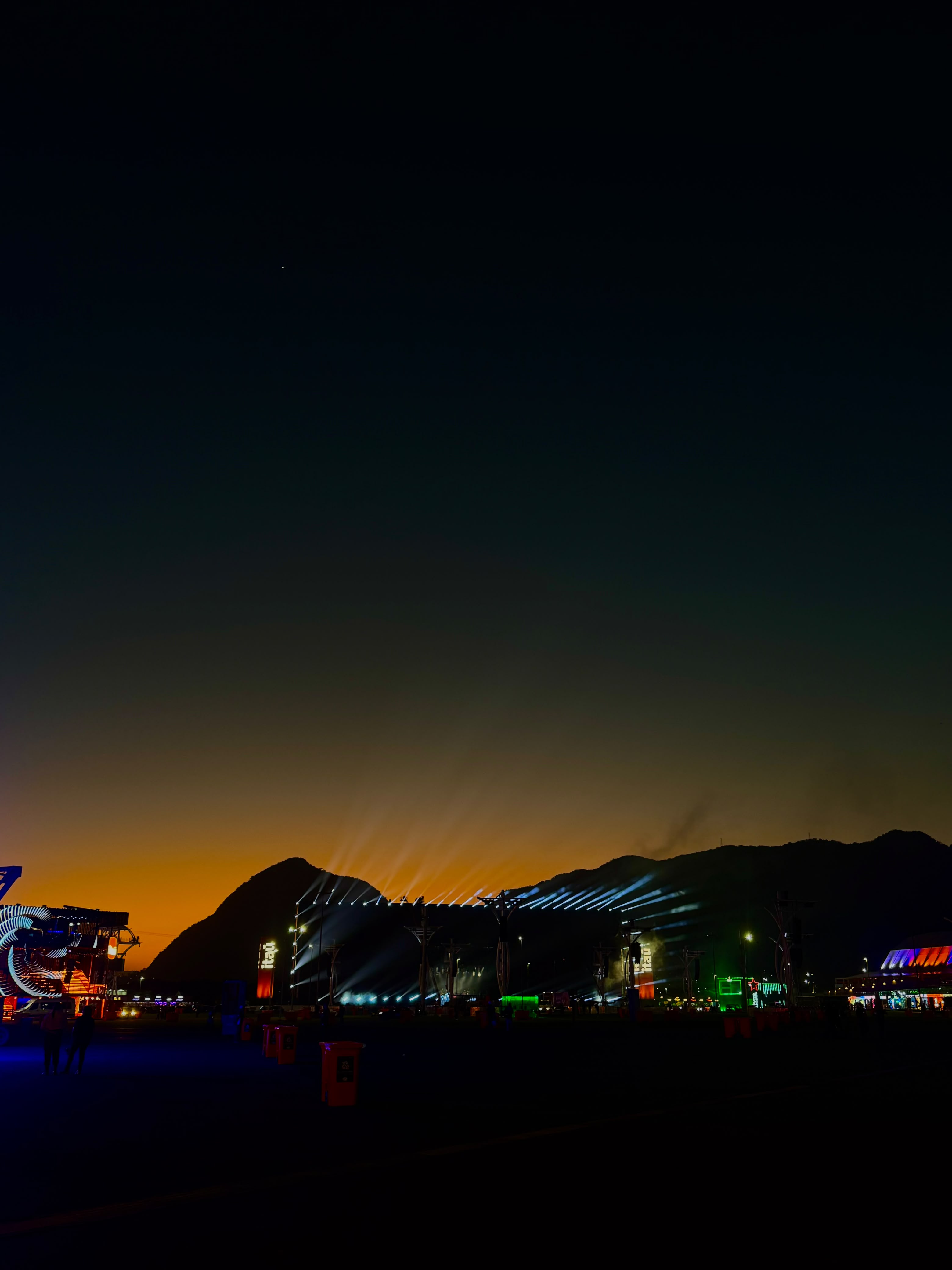 Palco de festival iluminado ao anoitecer, com feixes de luz apontando para o céu e montanhas em silhueta ao fundo.