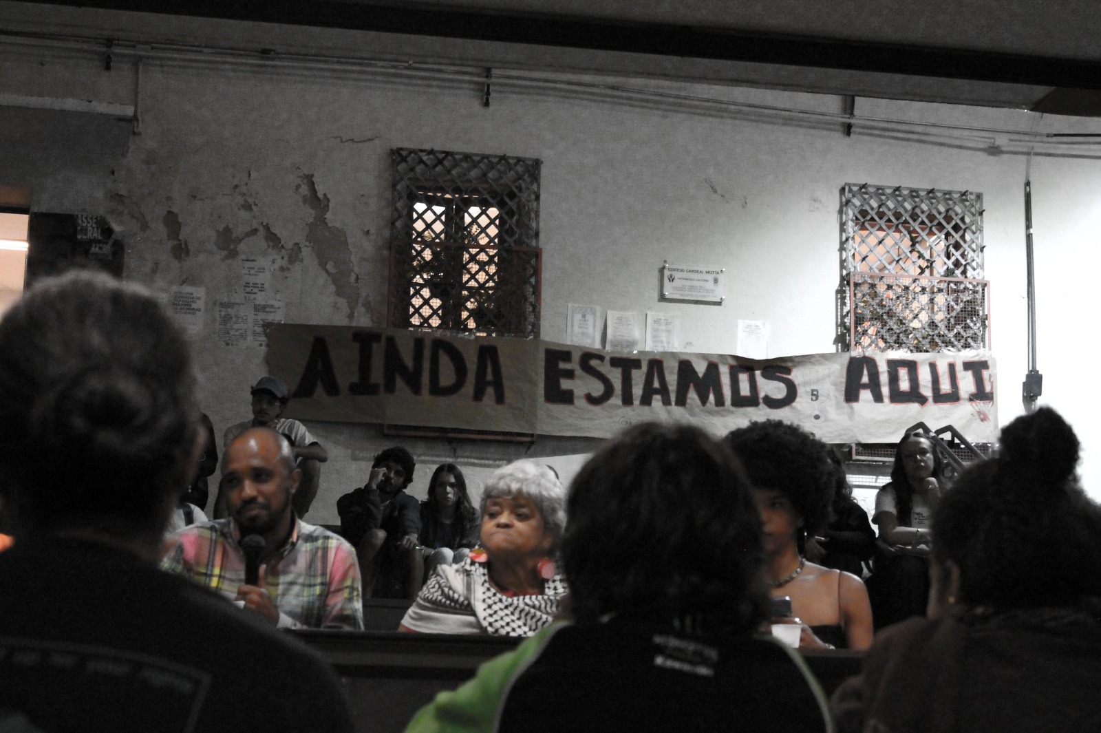 A imagem acima mostra três palestrantes em uma mesa durante uma fala na Pontifícia Universidade Católica de São Paulo, com uma faixa escrita "Ainda estamos aqui"