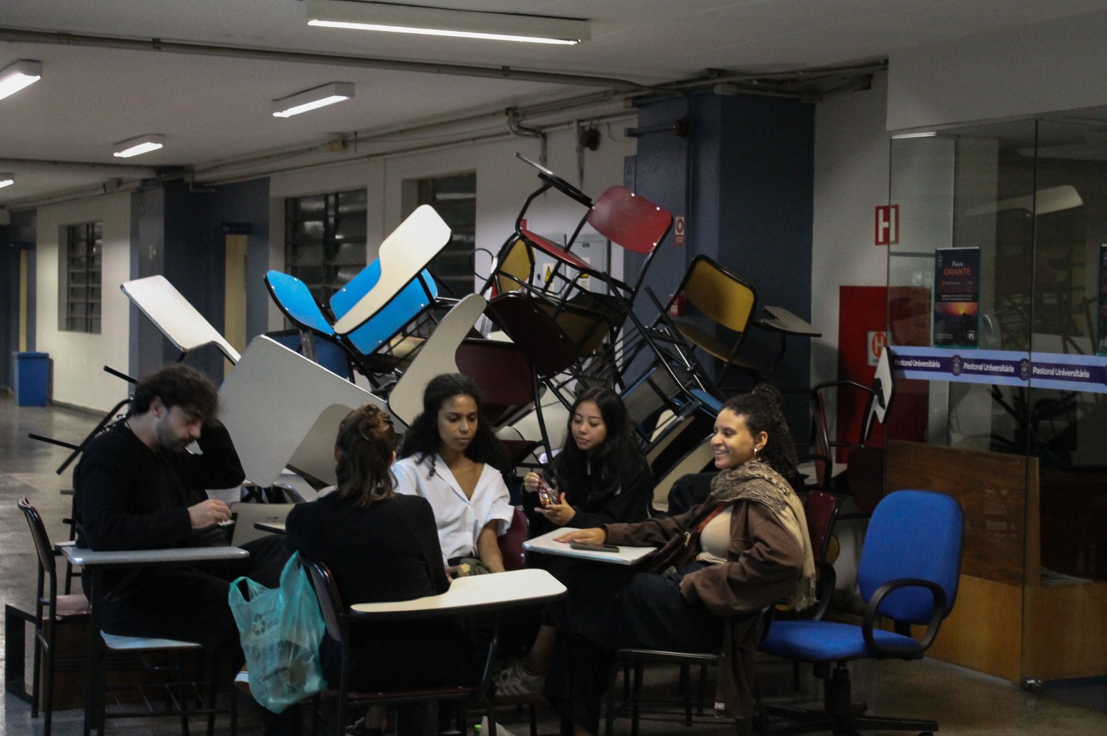 A imagem acima contém estudantes reunidos sentados em carteiras em frente a um bloqueio de cadeiras e mesas empilhadas em um corredor da Pontifícia Universidade Católica de São Paulo