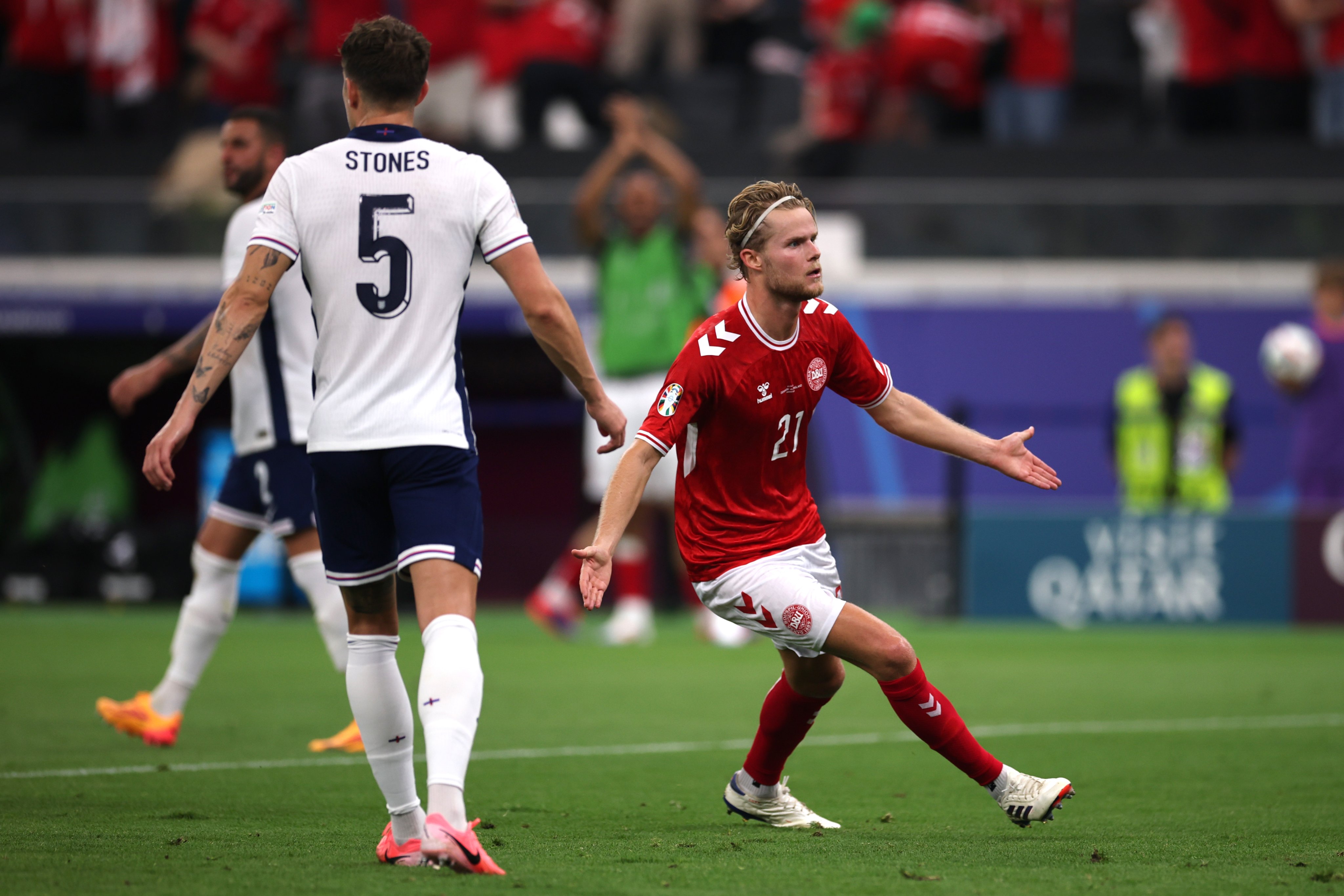 O jogador dinamarquês, Hjulmand, comemora seu gol contra a Inglaterra, de braços abertos no meio do campo, enquanto um jogador adversário anda de costas ao seu lado.