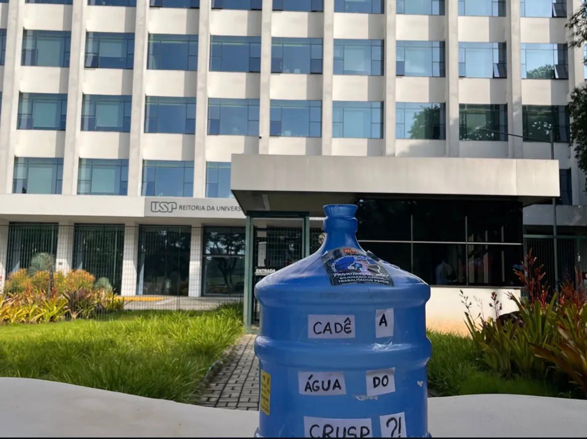 Galão de água com o escrito "Cadê a água do CRUSP?" em frente a reitoria. Reprodução:/ (@dceusp)
