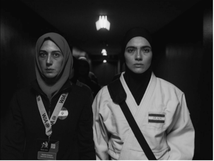 Na imagem em preto e branco está Maryam (Zar Amir) de pé à esquerda, usando um quimono preto e um hijab cinza. Na direita está Leila Hosseine (Arienne Mandi), de quimono branco e hijab preto. Ambas as mulheres olham para a frente com feições sérias.