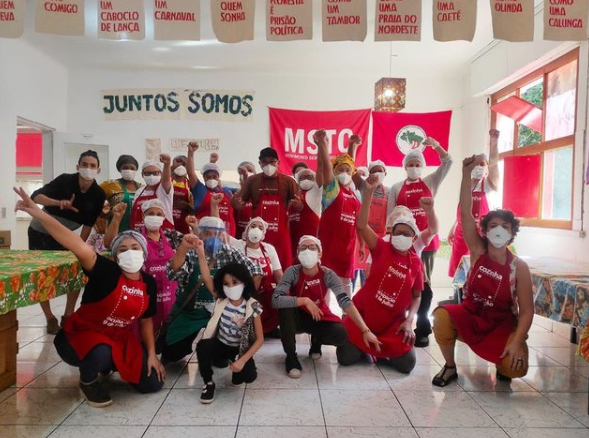 Na cozinha da ocupação 9 de julho, militantes da campanha Lute Como Quem Cuida posam com os punhos pra cima, em sinal de resistência. Atrás dos mesmos, existem duas bandeiras, sendo uma do Movimento Sem-Teto do Centro e outra do Movimento dos Trabalhadores Rurais Sem-Terra.