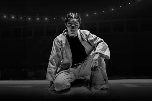 Na imagem em preto e branco está Leila Hosseine (Arienne Mandi) ajoelhada no meio de um tatame. Ela usa um quimono branco e seus cabelos estão trançados e a mostra.