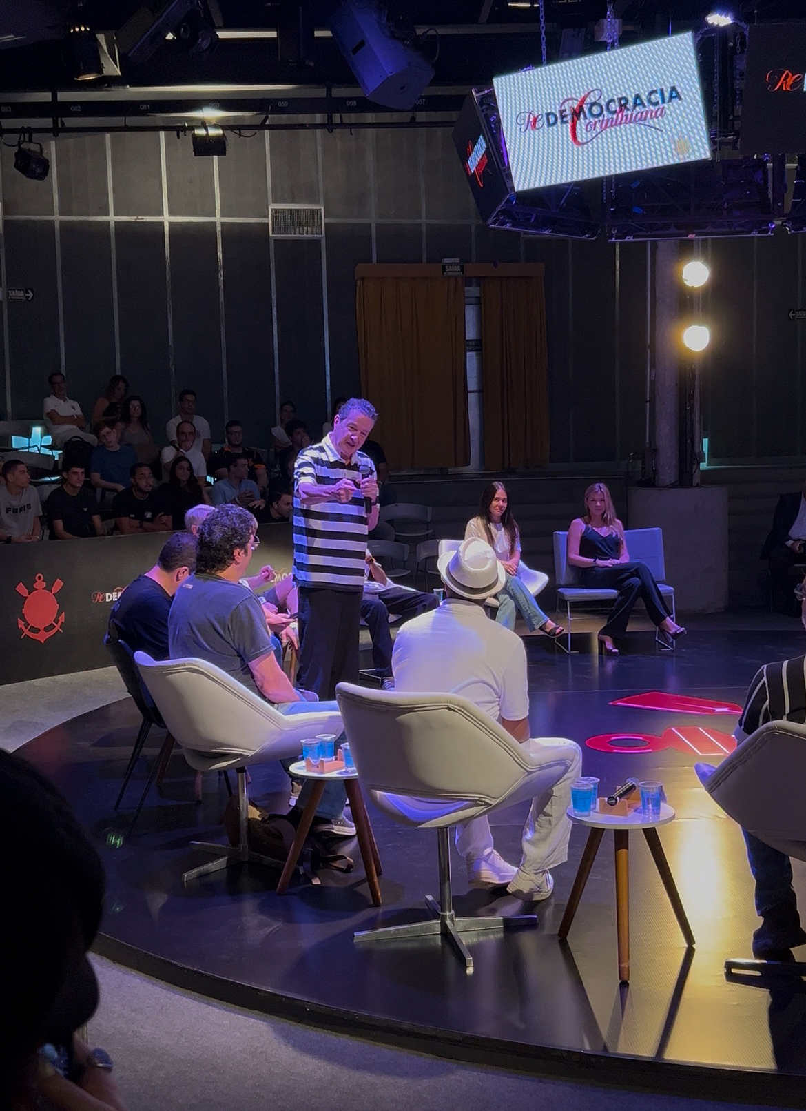 A imagem mostra Juca Kfouri em pé com o microfone na mão. Em volta, estão os participantes da mesa sentados