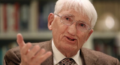 Fotografia em close-up de Jürgen Habermas, um homem idoso com cabelos brancos curtos e óculos de grau de armação fina. Ele veste um paletó de tweed marrom sobre uma camisa branca e gravata xadrez. Ele está com a mão direita levemente levantada em um gesto expressivo, como se estivesse explicando um conceito. O fundo está desfocado, sugerindo um ambiente de biblioteca ou escritório com estantes de livros.