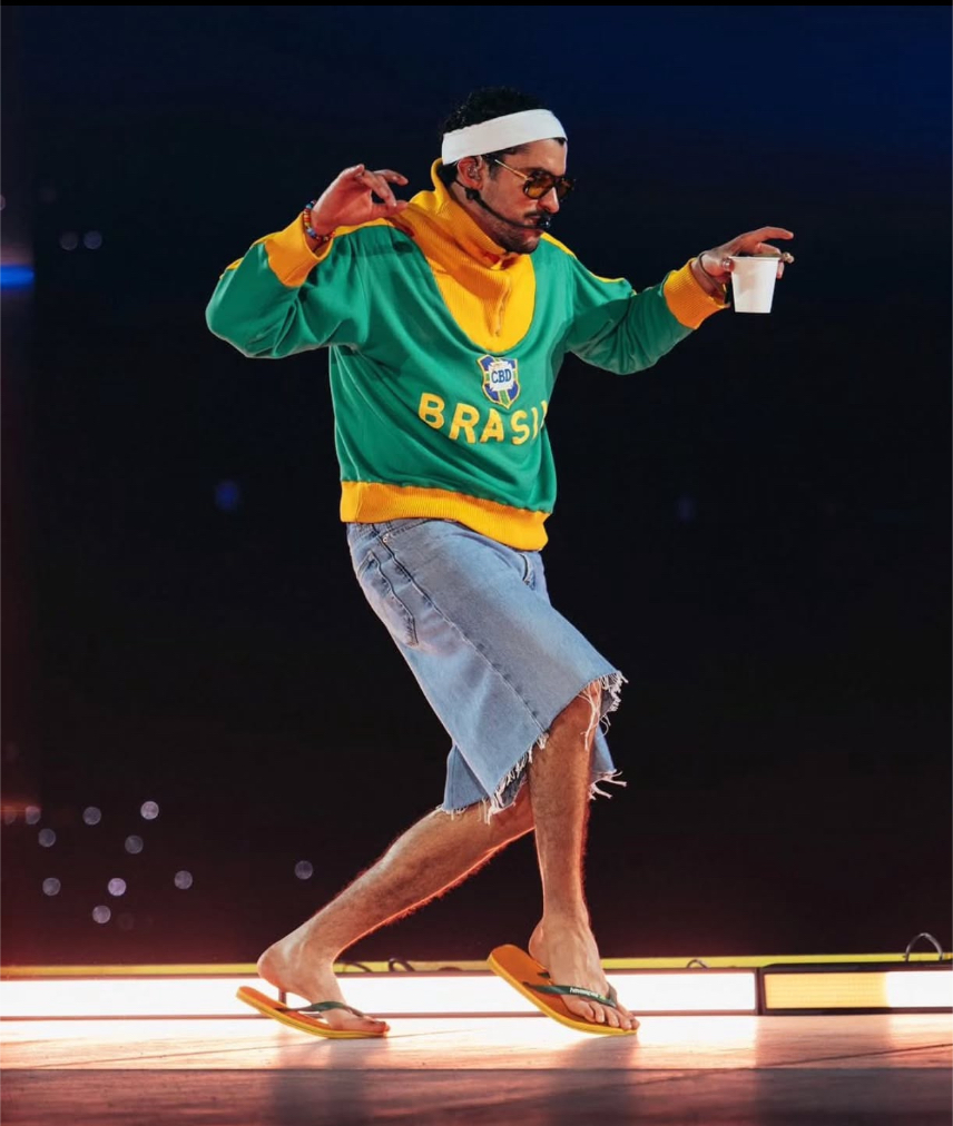 O cantor Bad Bunny é retratado de corpo inteiro em um palco iluminado, capturado em uma pose de dança dinâmica. Ele veste um moletom de gola alta nas cores verde e amarelo com a palavra "BRASIL" e um escudo retrô da CBD no peito. Complementando o visual, ele usa bermuda jeans de corte irregular (oversized), óculos de sol escuros e uma faixa branca na cabeça. Ele segura um copo descartável branco na mão esquerda enquanto equilibra o corpo de forma descontraída, com o fundo do palco escuro e luzes desfocadas ao longe.