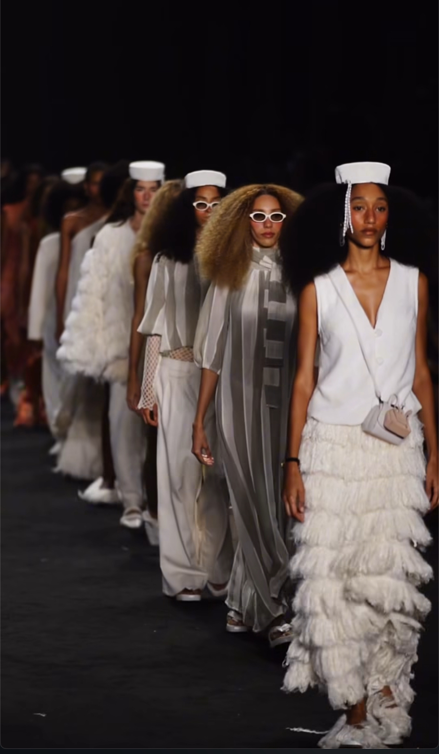 Pessoas desfilam em fila única sobre uma passarela escura, vestindo looks predominantemente brancos e em tons claros. As peças têm tecidos leves, camadas, transparências e detalhes texturizados. Algumas usam óculos claros e acessórios discretos. O enquadramento mostra a sequência de looks em perspectiva, com iluminação focada nas roupas.
